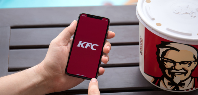 Logo do KFC em celular, ao lado de balde com comidas da empresa, representando uma procura por emprego no KFC.